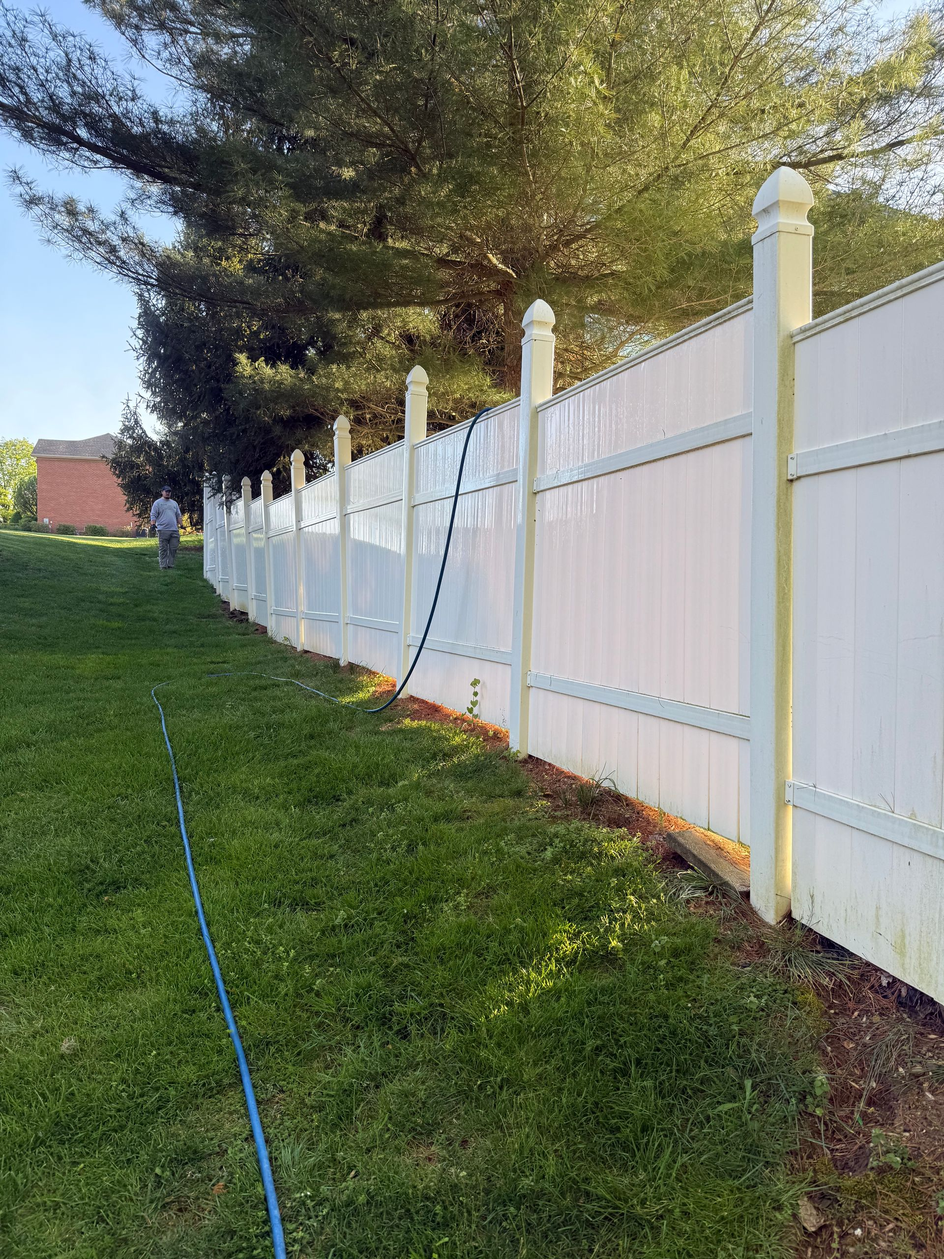 White vinyl fence along a grassy slope with trees and a garden hose in the foreground