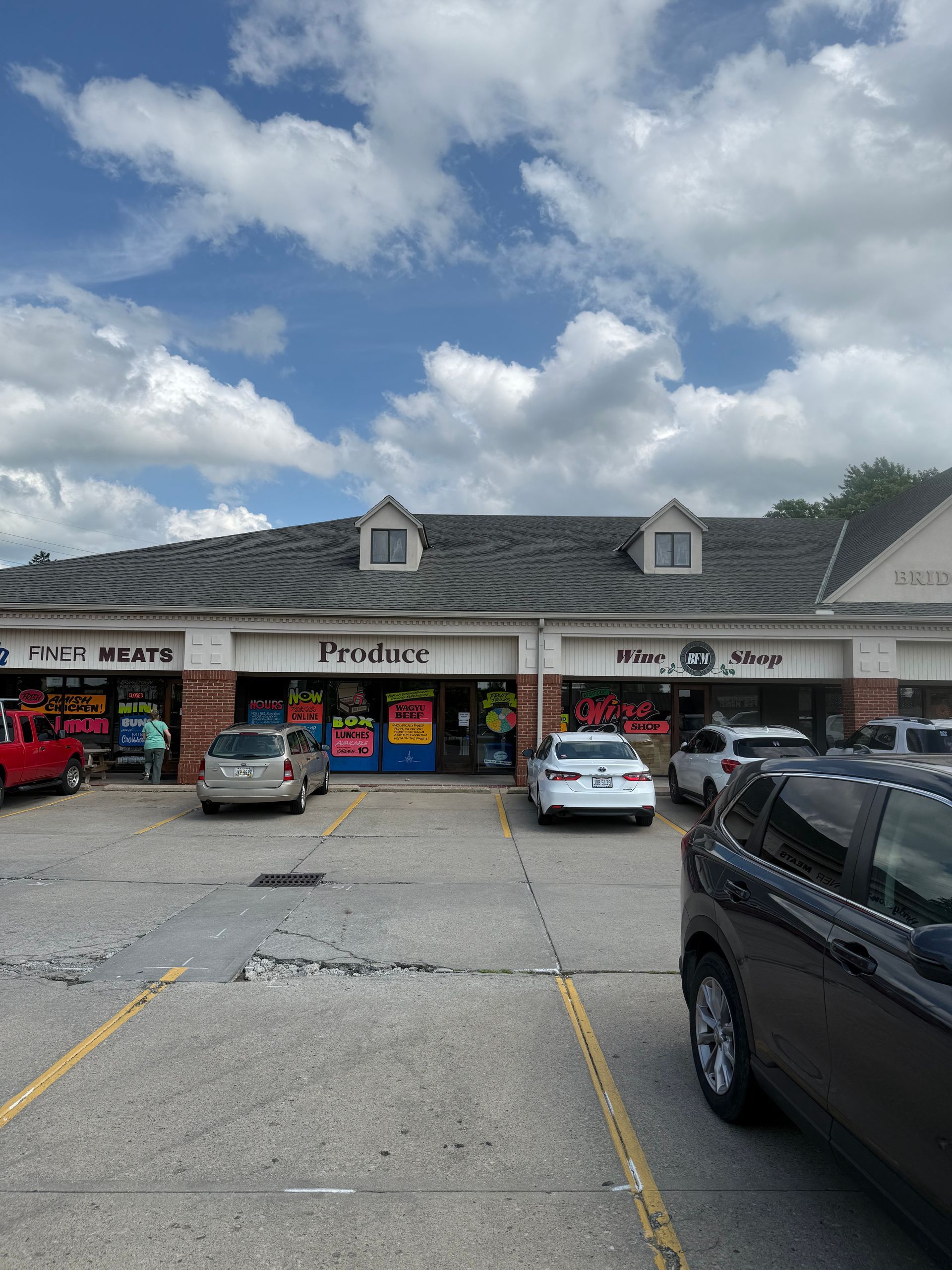Strip mall storefronts under a cloudy sky, with parked cars in front.
