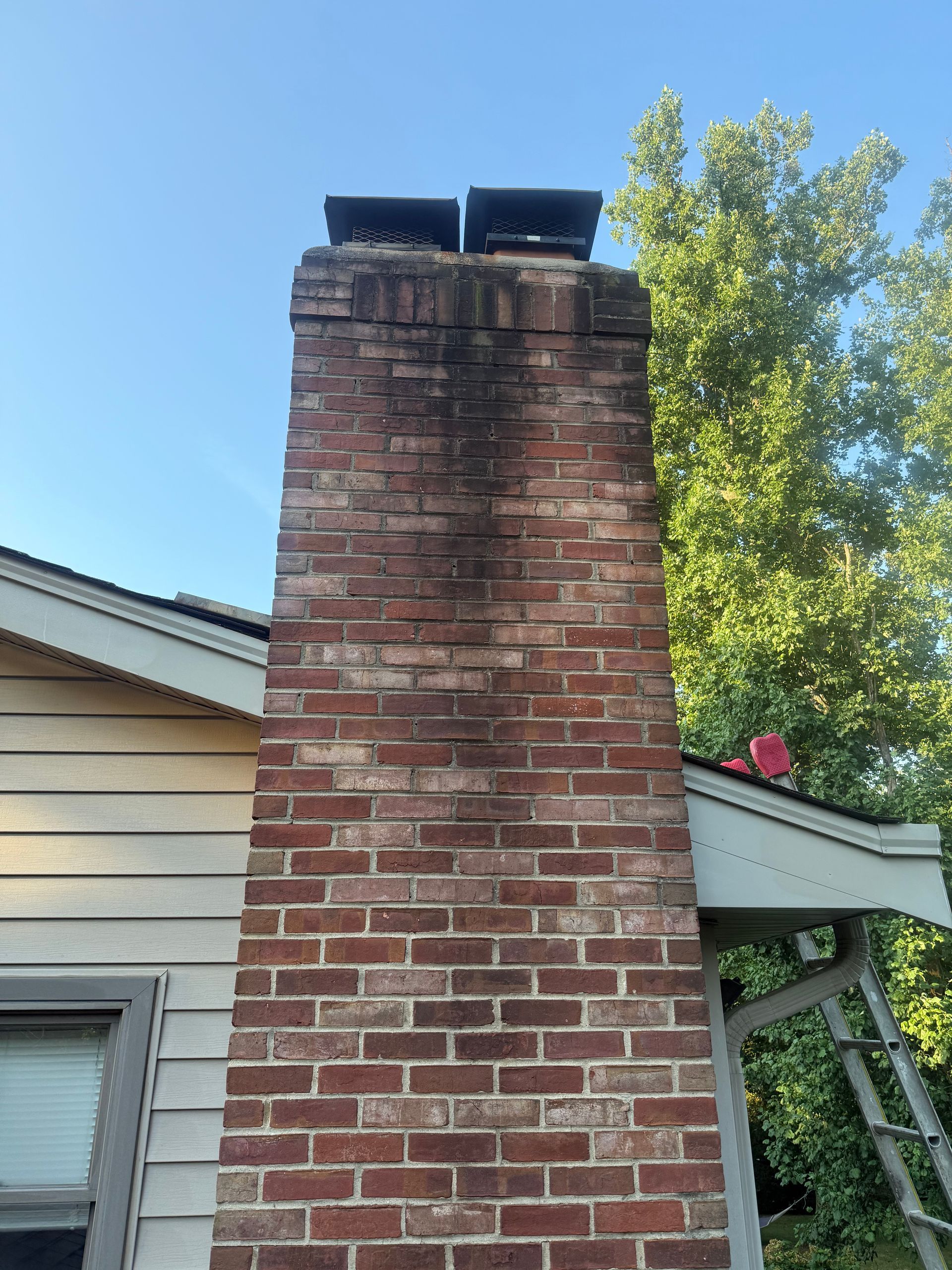 Tall brick chimney with black cap beside a house roof, framed against a blue sky and trees.