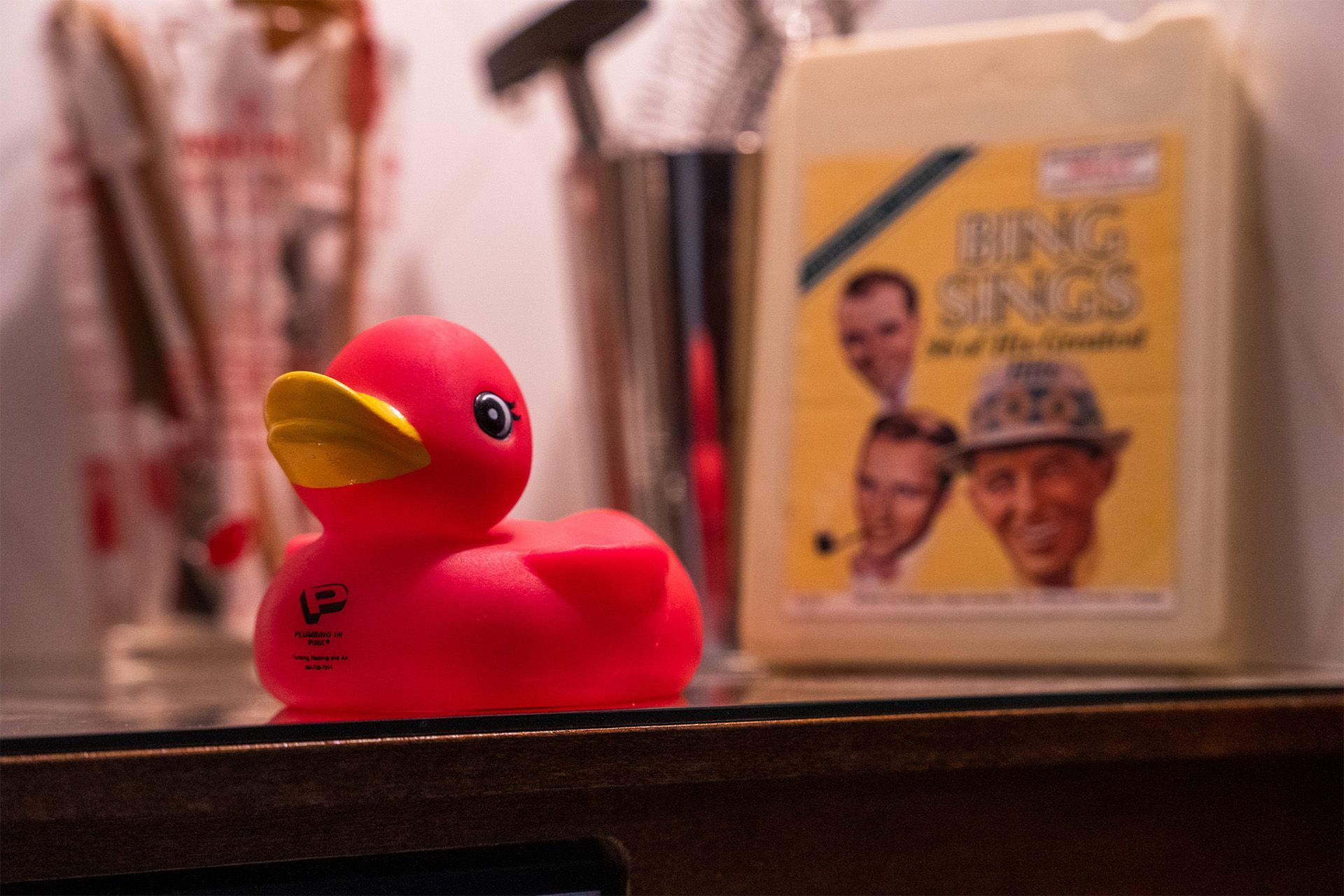 Pink rubber duck with yellow beak on a shelf, with a blurred book or magazine in the background