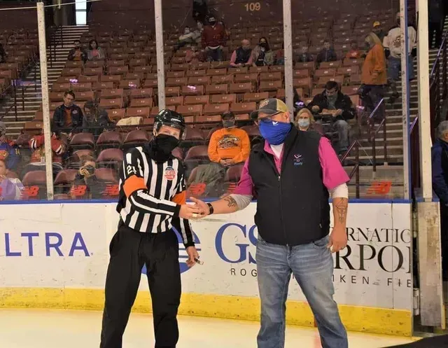 Ice rink referee in striped jersey shakes hands with a man in a blue mask near the boards.