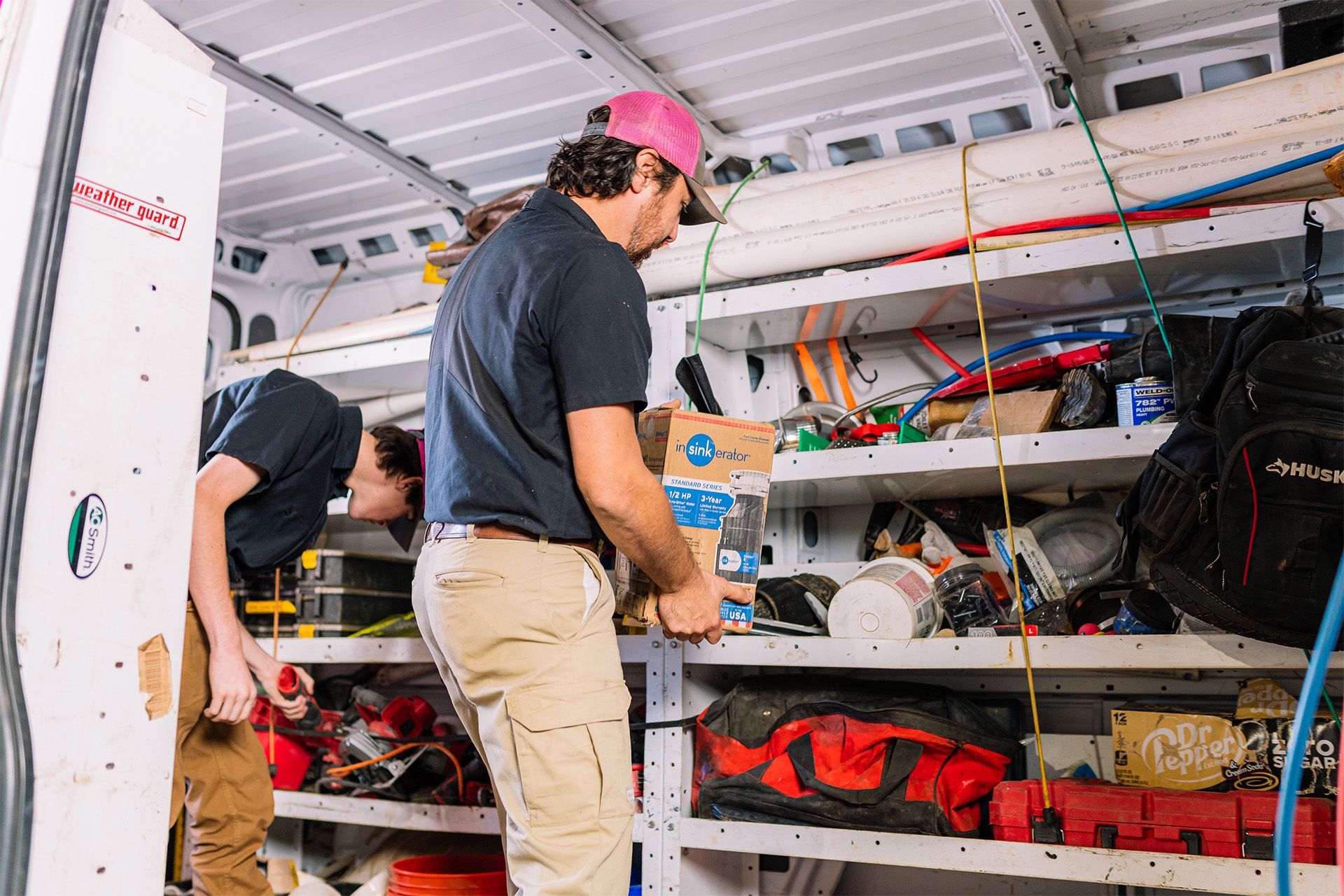 Two people organizing equipment on shelves inside a white van with tools and gear scattered around