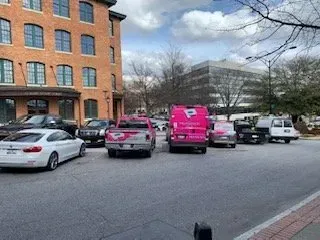 Parking lot with several cars, including a bright pink van, beside a brick building on a cloudy day