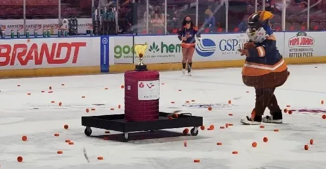 Mascot in a hockey arena beside a red canister on a cart, surrounded by scattered orange balls on the ice