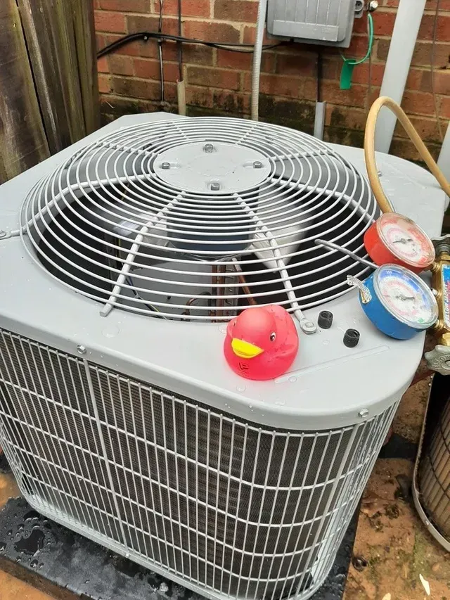 Outdoor HVAC condenser unit with white top grille, red rubber duck, and gauges on the side beside a brick wall