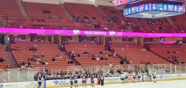 Indoor hockey arena with orange seats and a few spectators, viewed from the stands above the rink.