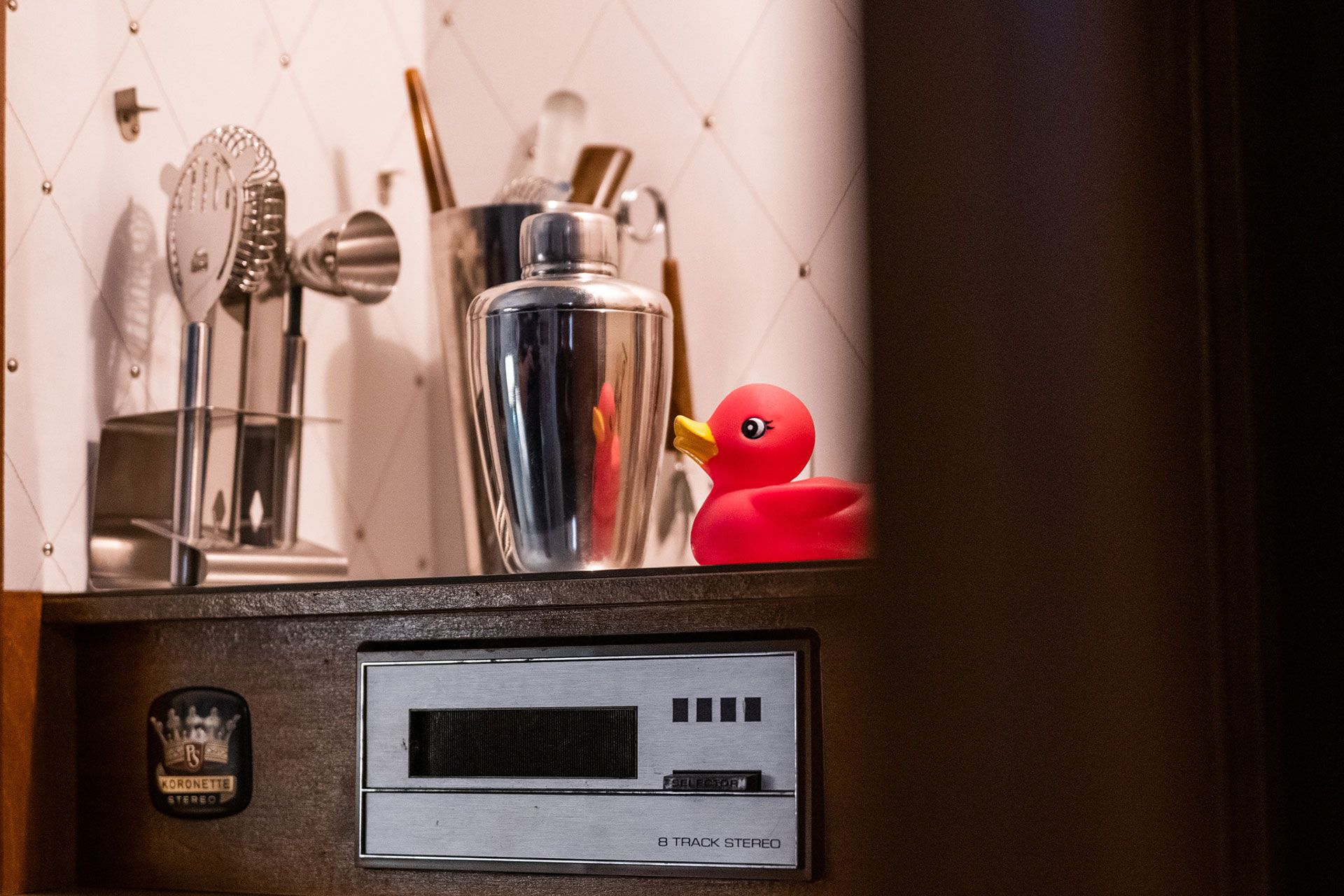 Shelf with cocktail tools, a silver shaker, and a red rubber duck above a vintage radio.