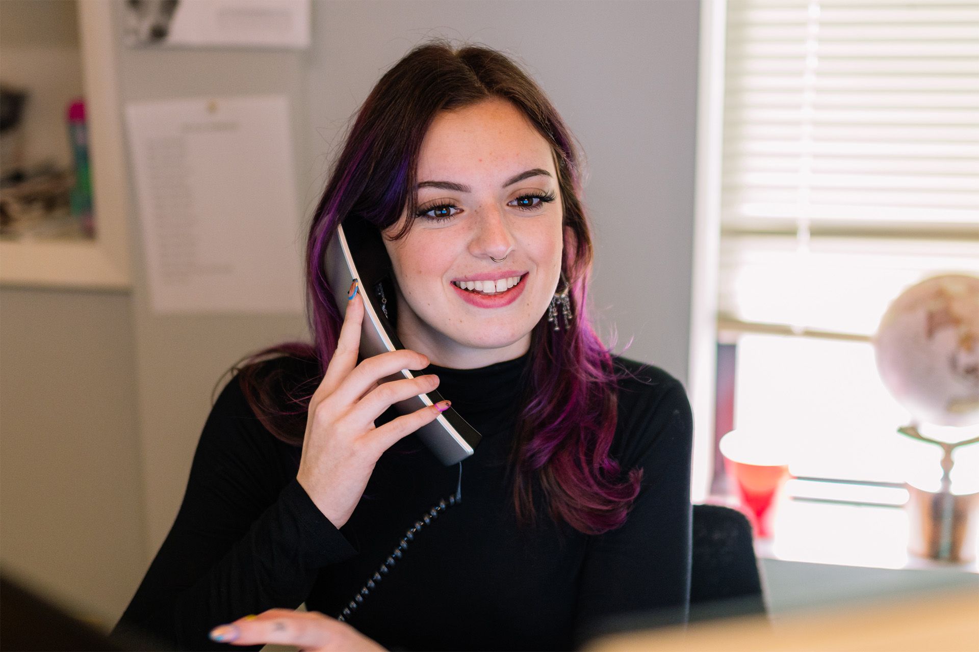 Woman in a black sweater smiling while talking on a phone in a bright office.