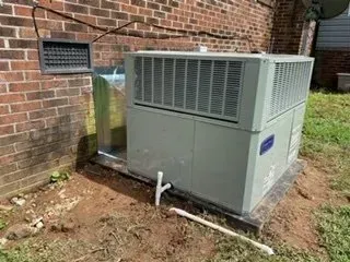 Outdoor HVAC unit beside a brick building, with exposed metal piping and dirt around the base