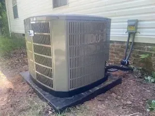 Outdoor HVAC condenser unit beside a house on a concrete pad, with pipes and utility lines visible.