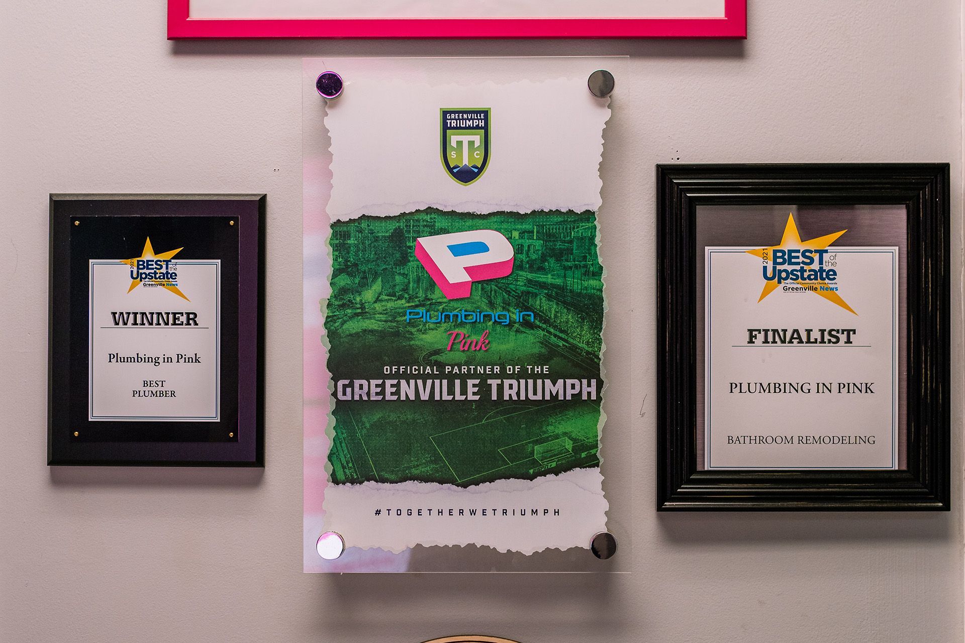 Three framed awards and a Greenville Triumph poster hanging on a wall.