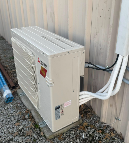 Mitsubishi Electric heat pump unit mounted on a wooden base, beside a building. White pipes lead to the electrical box.