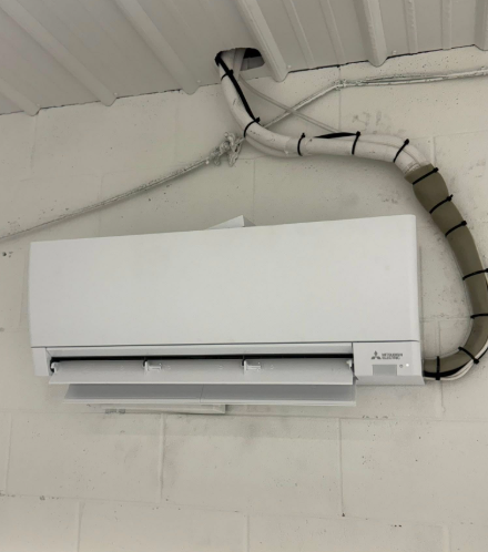 White Mitsubishi air conditioner mounted on a white brick wall, with insulated lines running to the ceiling.