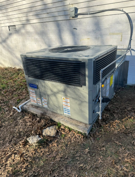 Outdoor air conditioning unit, gray metal, on wooden base next to a white house, surrounded by brown leaves.