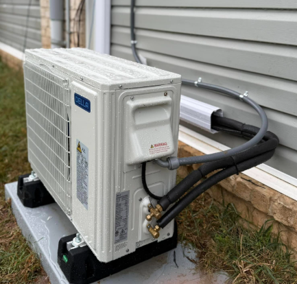 Outdoor heat pump unit on a gray platform next to a light gray wall; black tubing and white conduit attached.