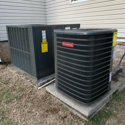Two gray HVAC units on concrete pads next to a beige house, Goodman brand visible.