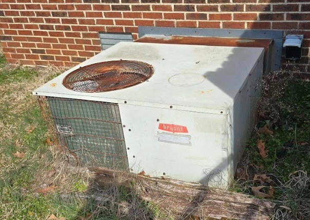 An old, rusty air conditioning unit against a brick wall and overgrown grass.