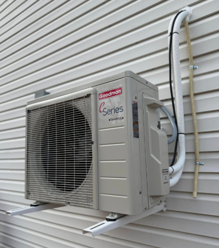 Wall-mounted Goodman air conditioner unit on a cream-colored siding wall. Unit has metal fan cover and attached tubing.