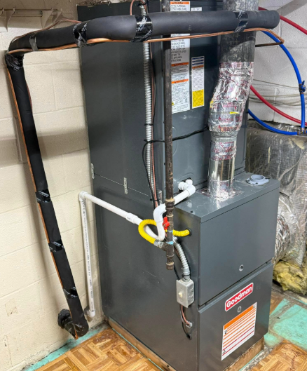 Gray Goodman furnace in a basement. Pipes and ducts with insulation are visible.