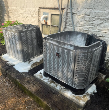Two outdoor air conditioning units on concrete pads, covered in foamy cleaner.