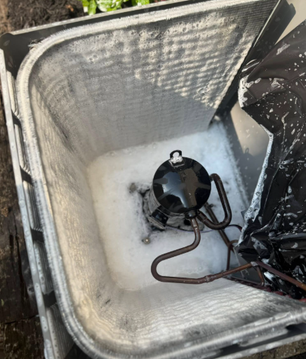 Air conditioner compressor submerged in soapy water inside a plastic bin for cleaning.