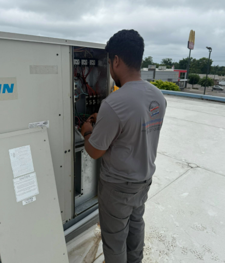 A person in gray shirt working on electrical components of an air conditioning unit on a rooftop.