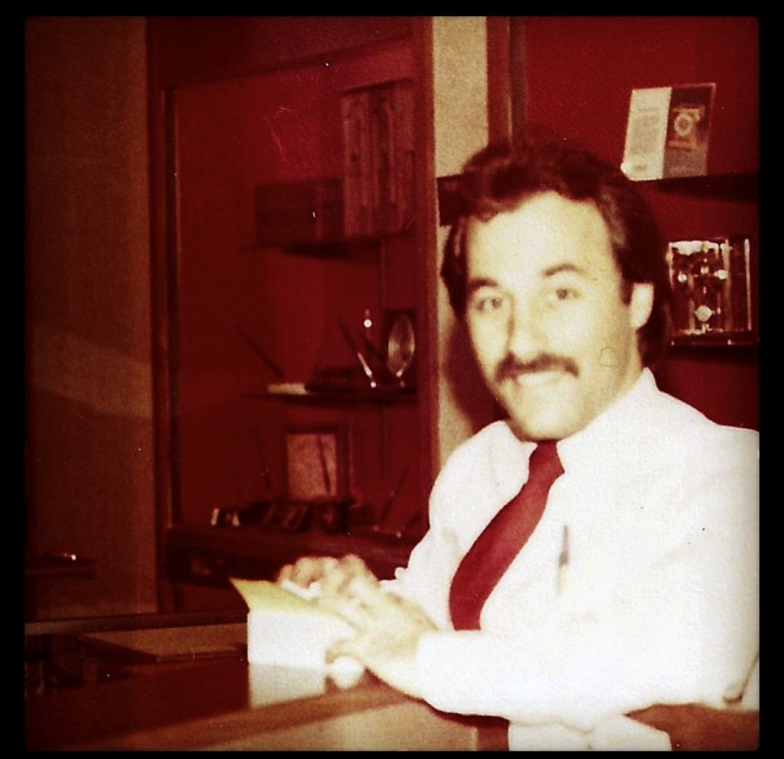 Man in white shirt and red tie seated at a desk, smiling in a warmly lit room with shelves behind him
