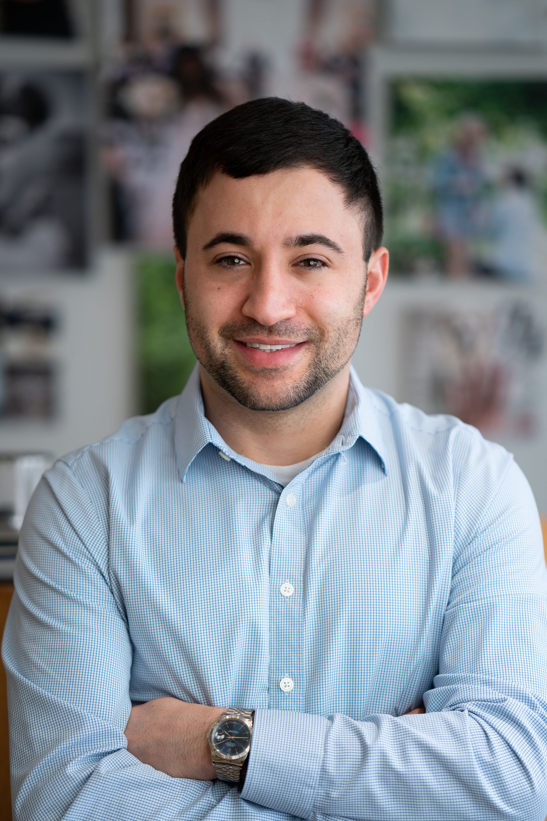 Smiling man in a light blue checkered shirt with arms crossed, standing indoors against a blurred wall.