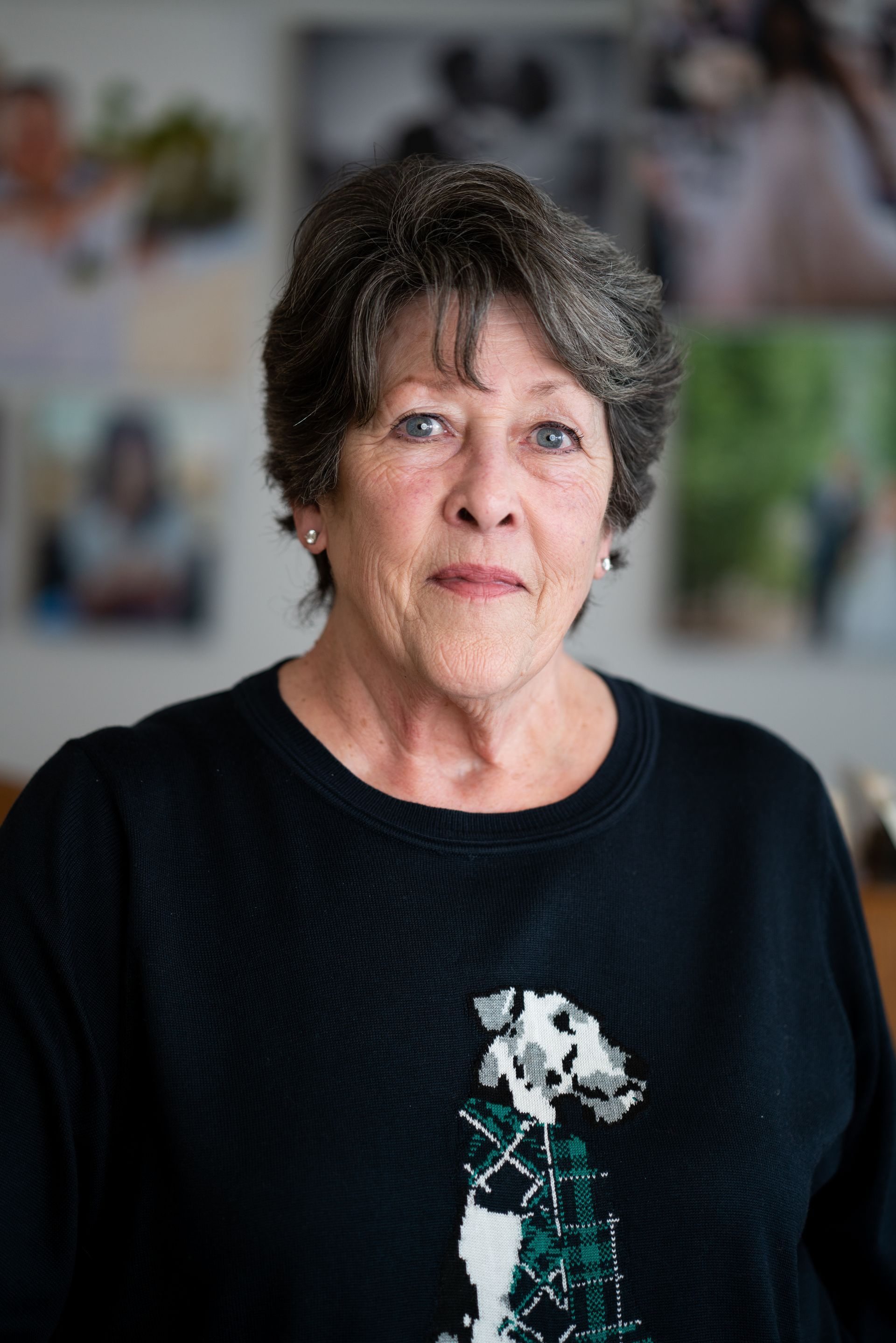 Woman in a black sweater standing indoors against a blurred background of photos and plants
