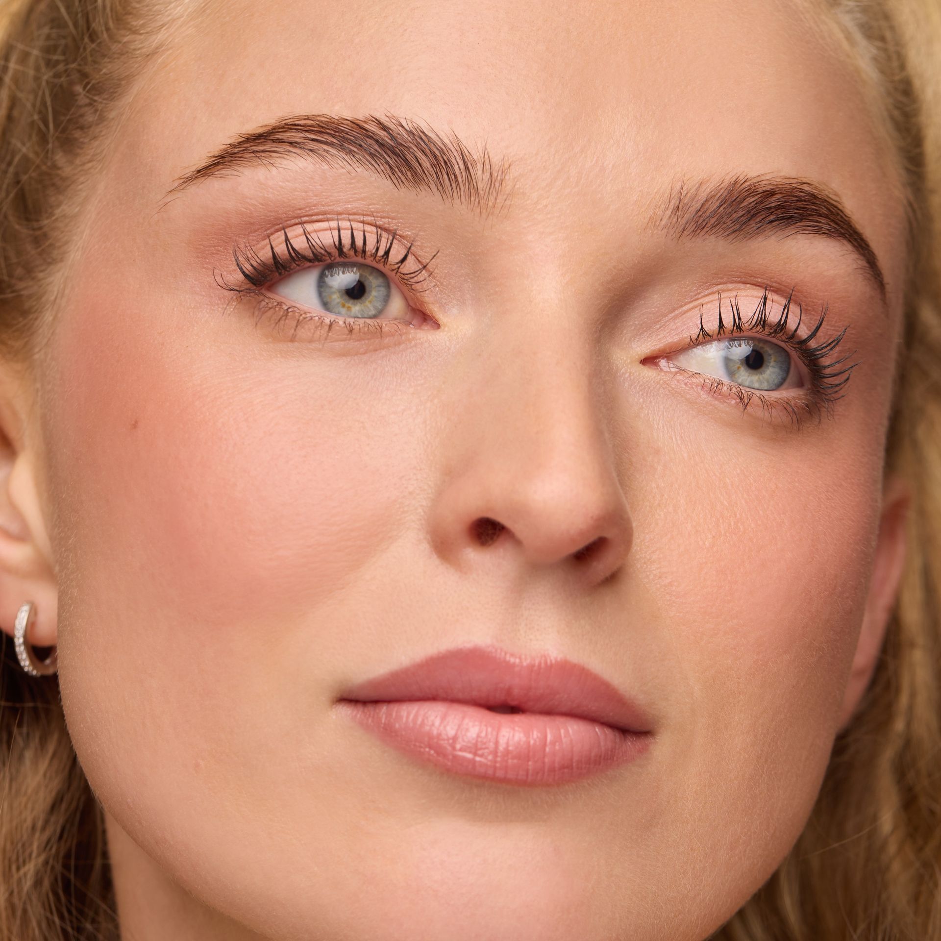 Close-up of a person with light eyes, defined lashes, natural-toned makeup, and a small silver hoop earring