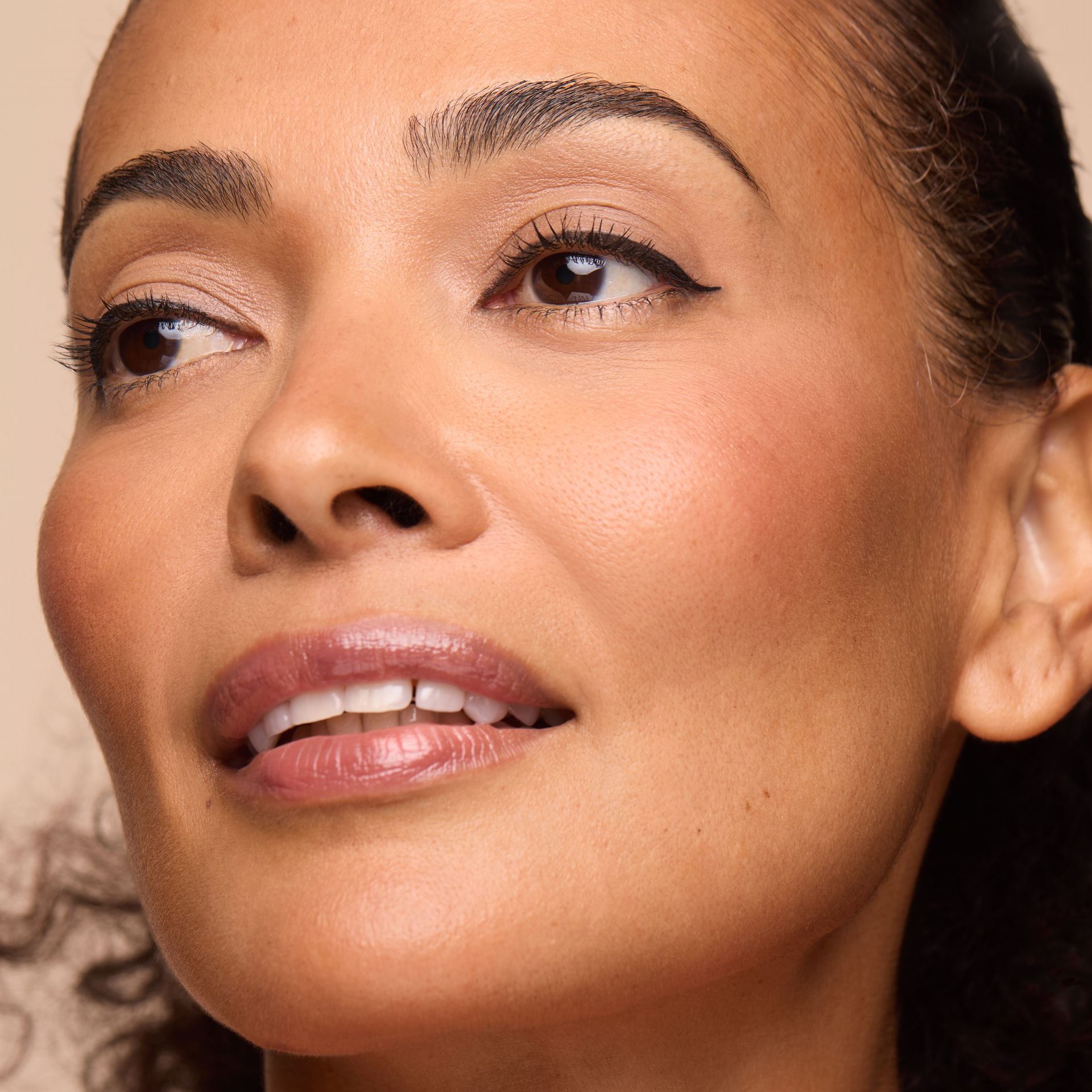 A person with a soft, natural makeup look, including subtle eyeliner and glossy mauve lips, posing against a beige backdrop