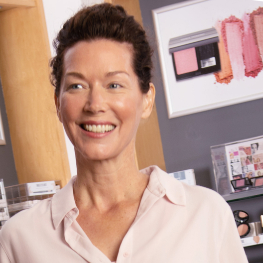 A smiling person in a light pink shirt standing in a retail store with makeup displays in the background