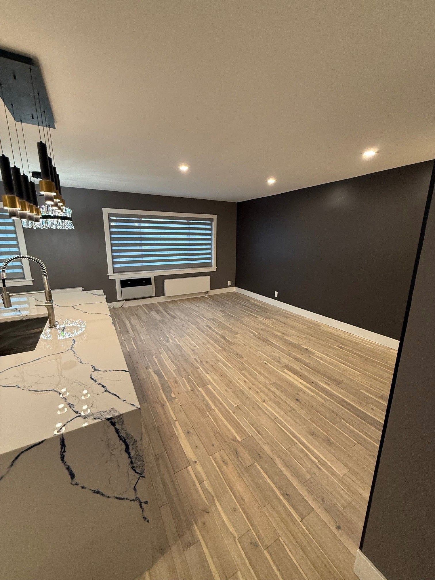 A modern, empty living room with light wood floors, dark accent walls, large windows, and an adjacent kitchen island.