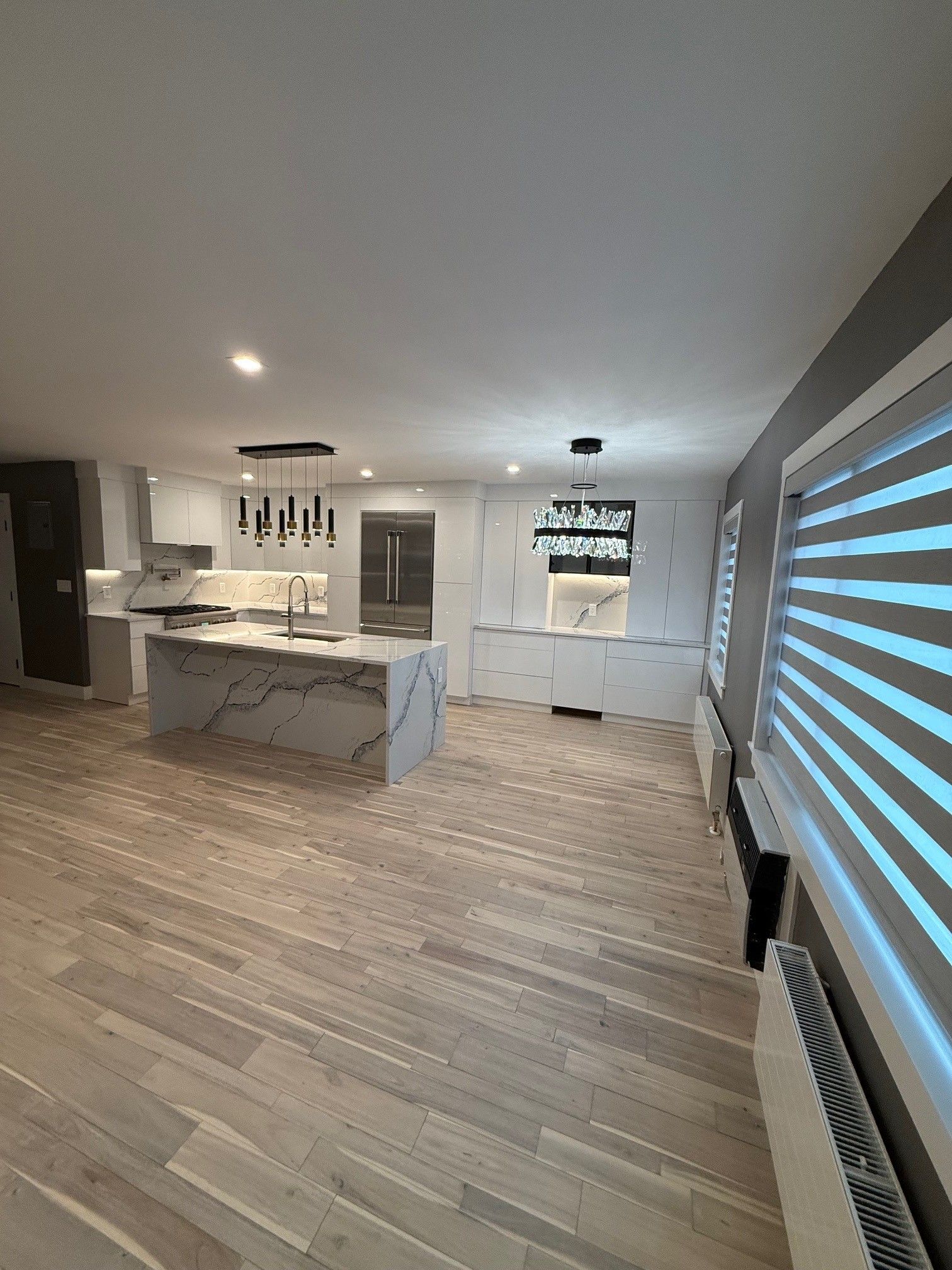 A bright modern kitchen with a white marble-topped island, light wooden floors, white cabinetry, and horizontal blinds.