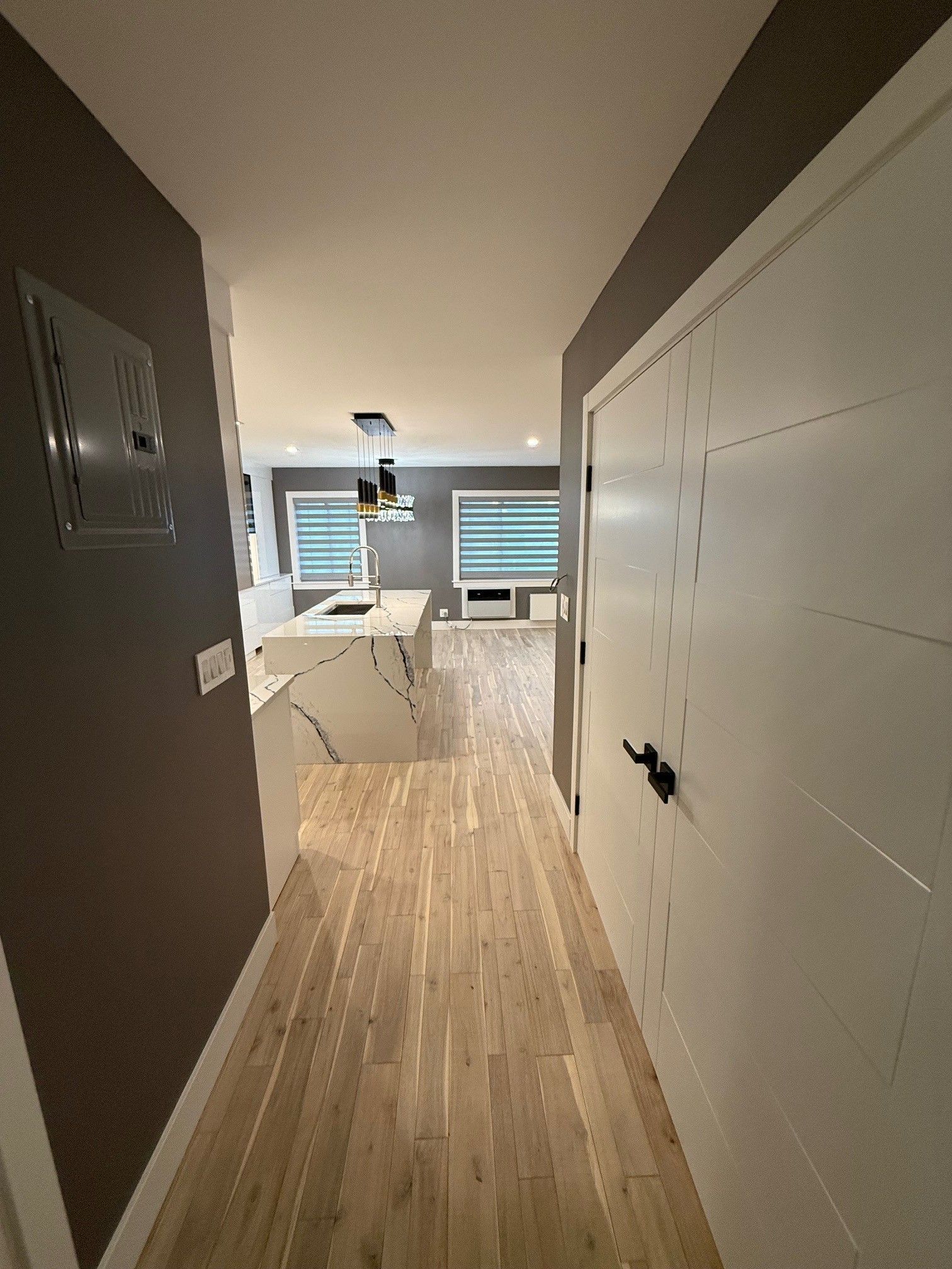 A hallway with light wood floors leading to an open-concept kitchen with a marble-topped island and white double doors.