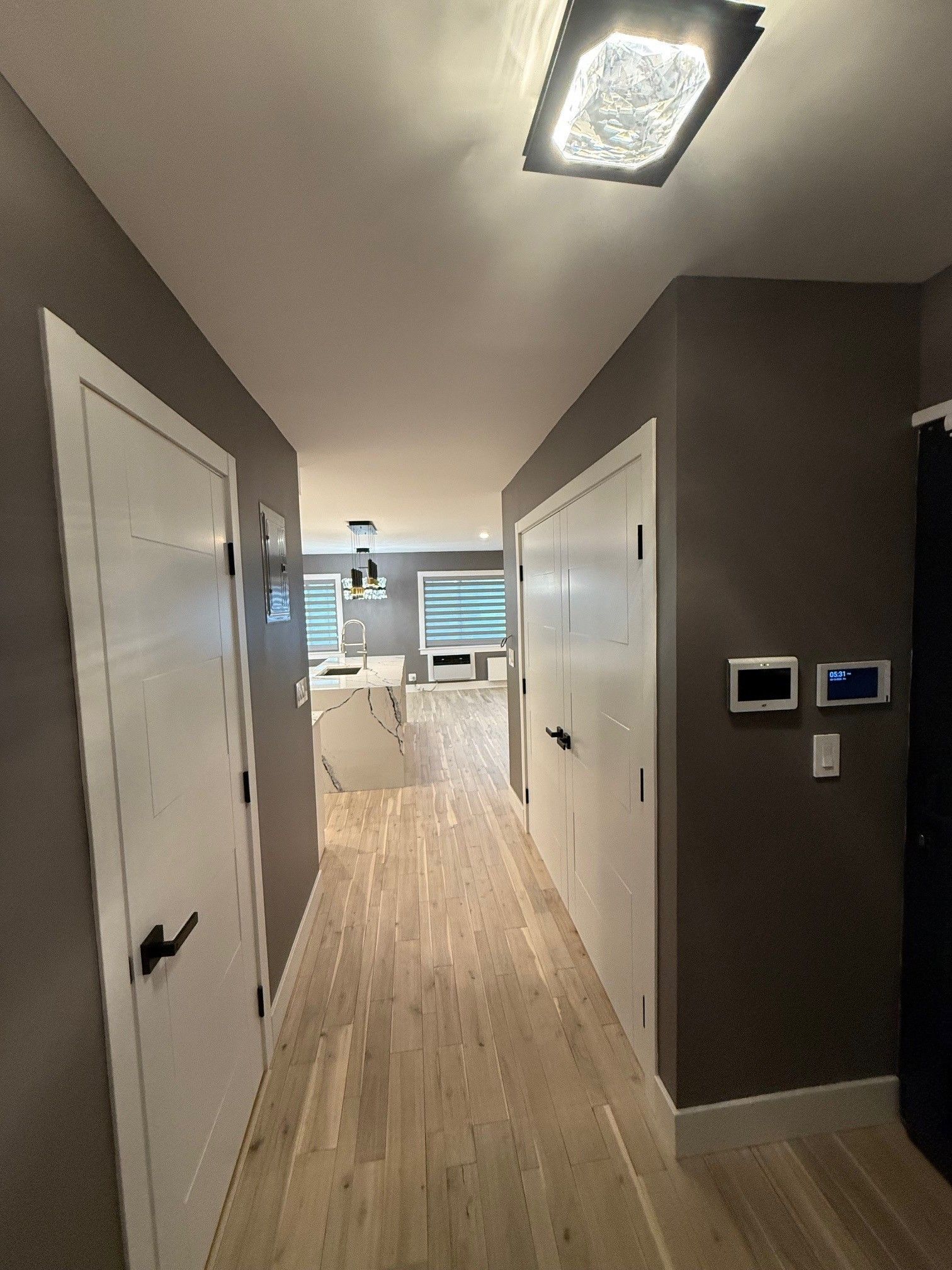 A hallway with light wood floors, grey walls, and white doors, leading toward a bright kitchen area with an island.