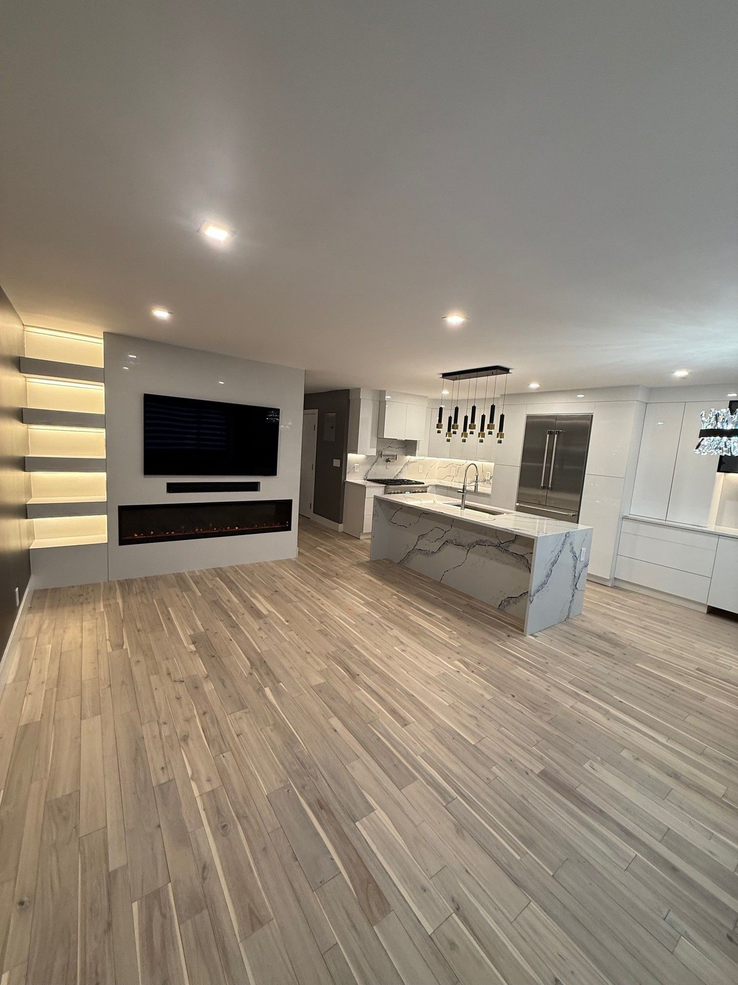 A wide view of a modern open-concept kitchen and living area with light wood flooring, a fireplace, and a marble island.