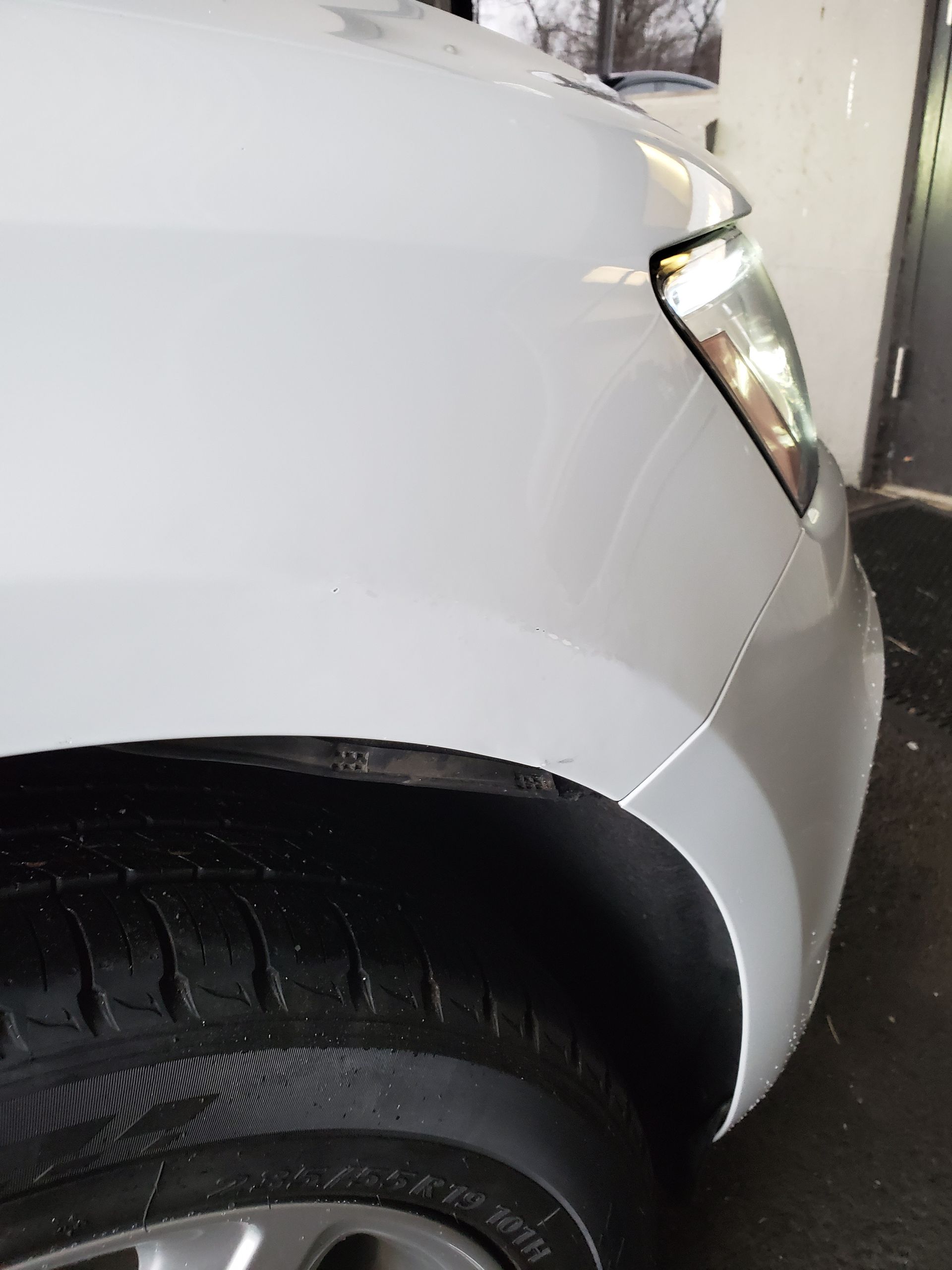 Close-up of a white car’s front fender and wheel near a wall, with headlights on.