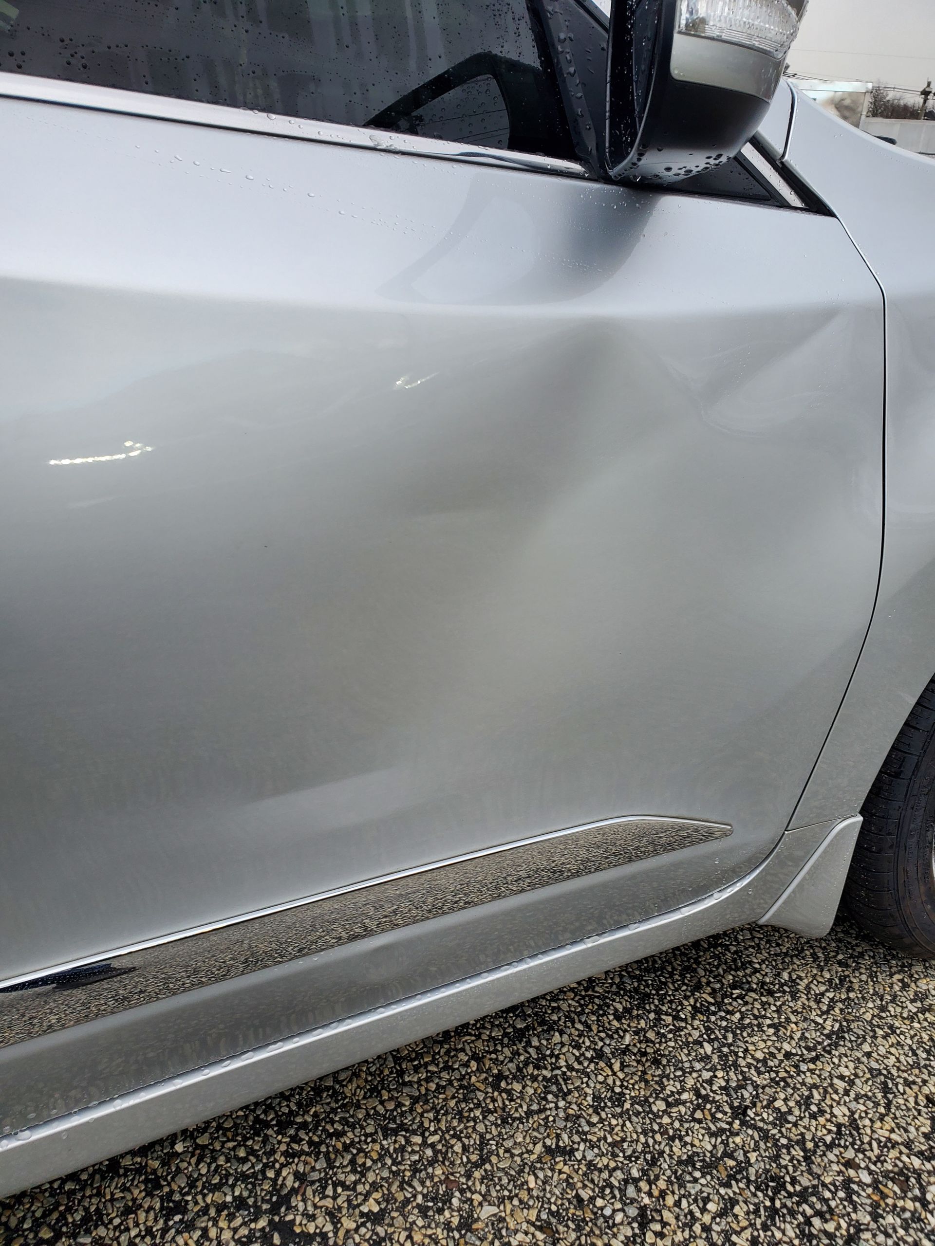Silver car door with a dent and scraped lower trim near the wheel