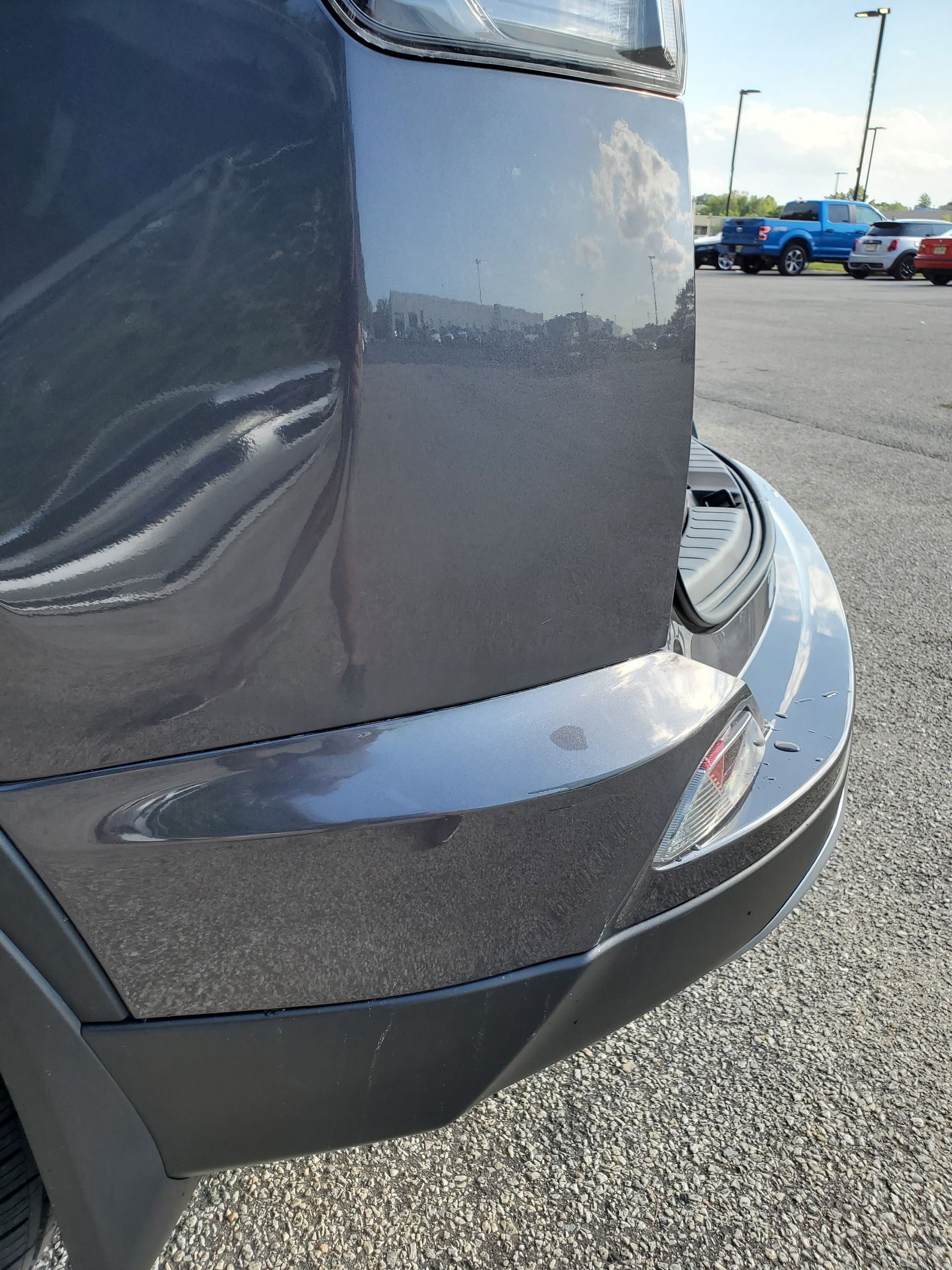 Close-up of a gray car’s rear bumper with a dent and scraped paint in a parking lot