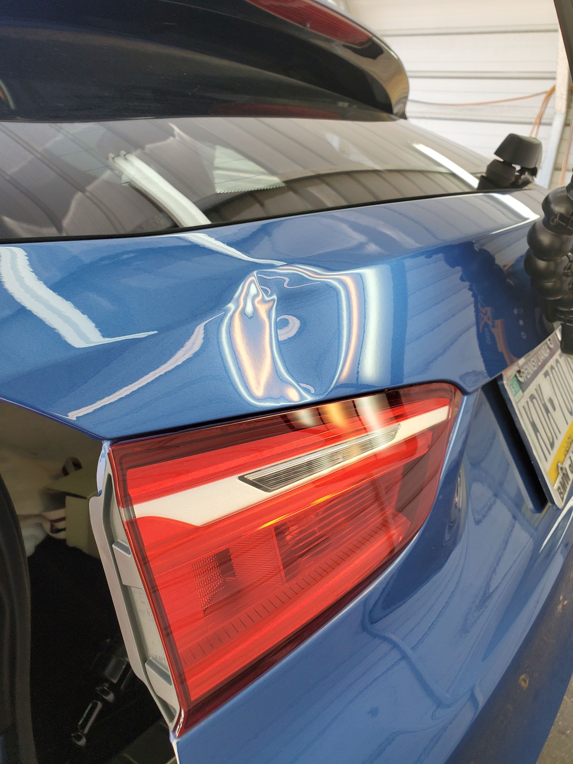 Blue car rear with dent above the taillight and a hanging license plate in a garage