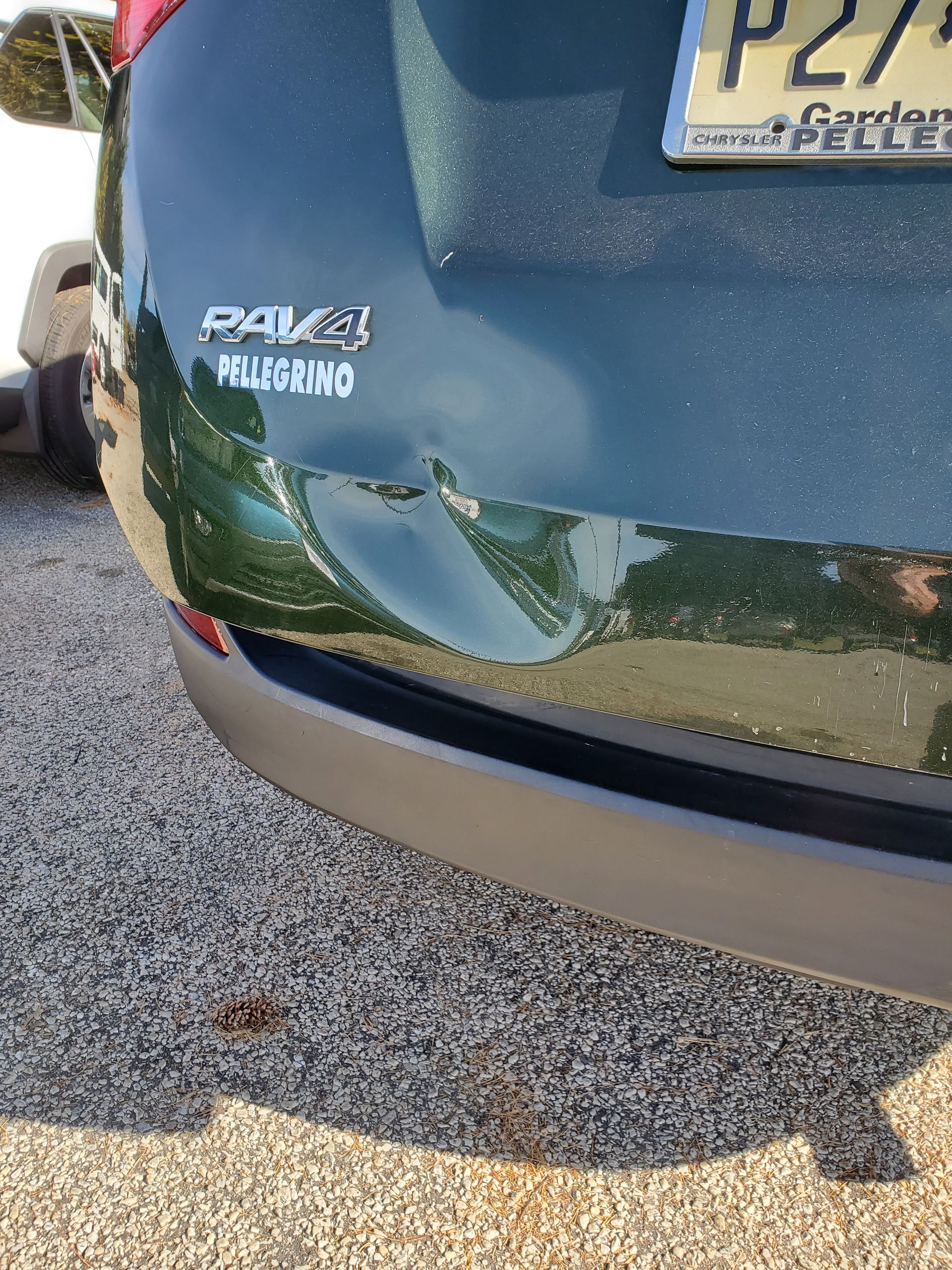 Rear of a dark blue car with a dented bumper and visible license plate, parked on gravel.