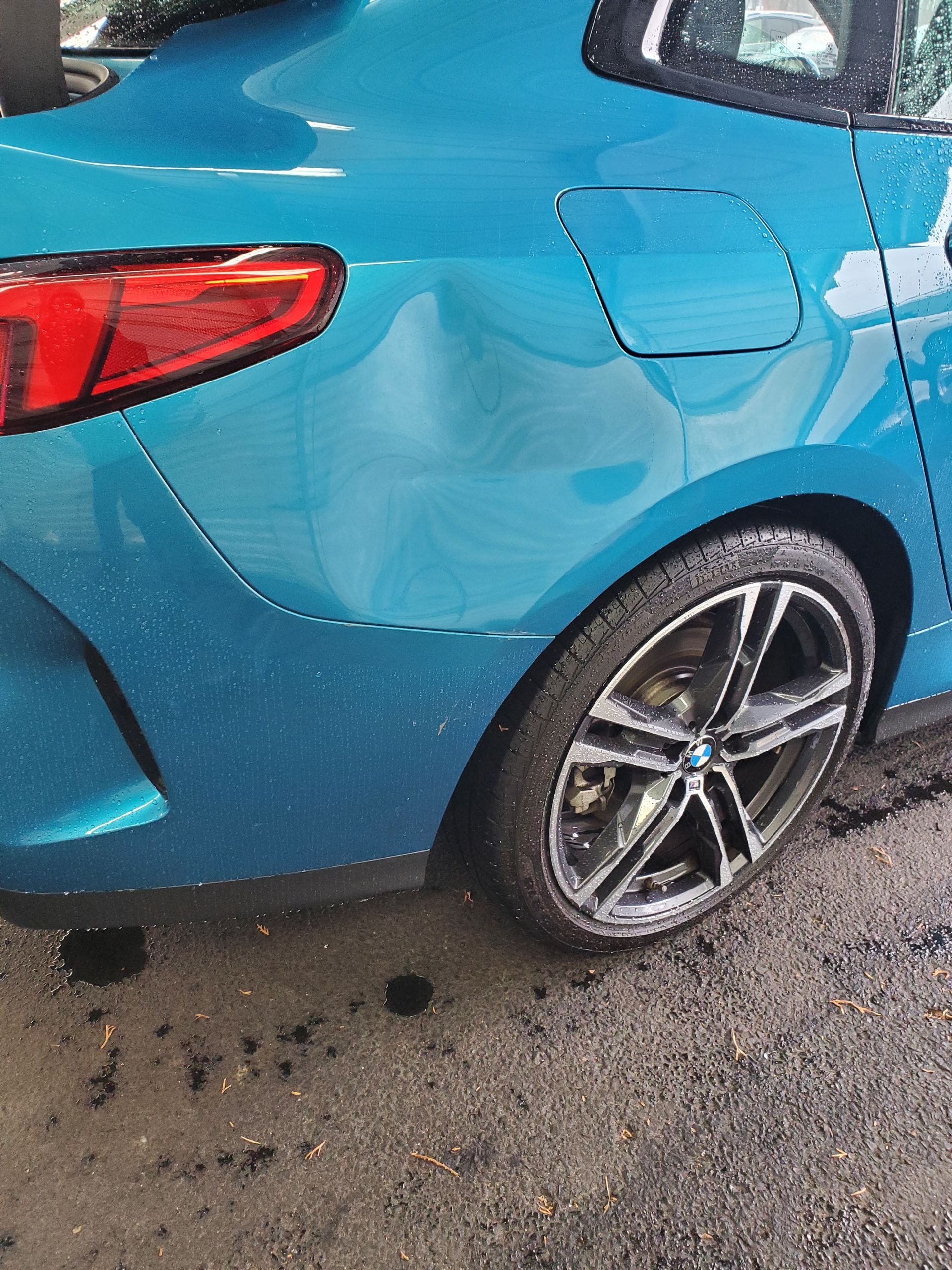 Blue car’s rear side with dented panel and wet pavement underneath