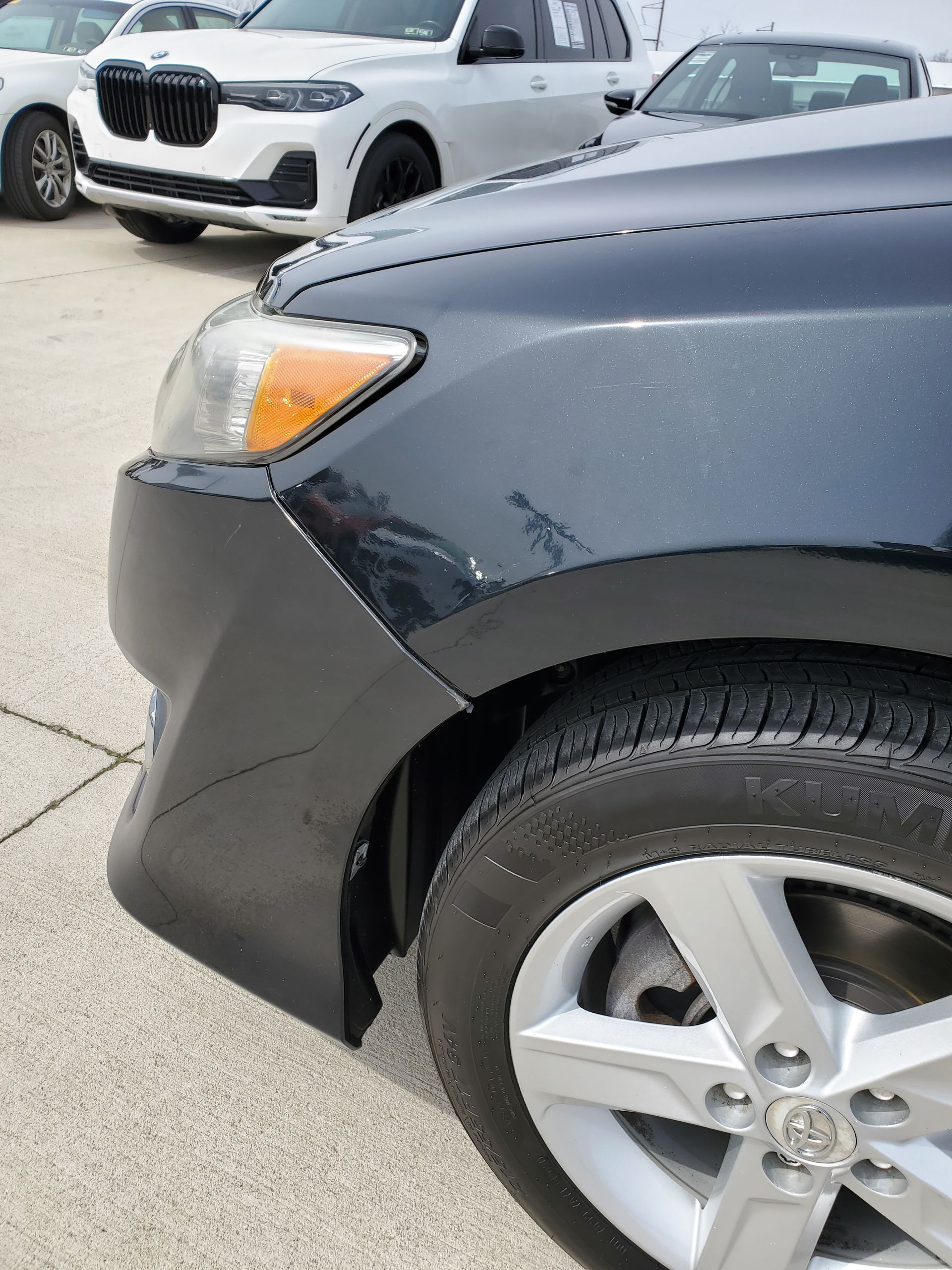 Close-up of black car’s front fender with a dent near the wheel, parked in a lot with a white SUV behind it.