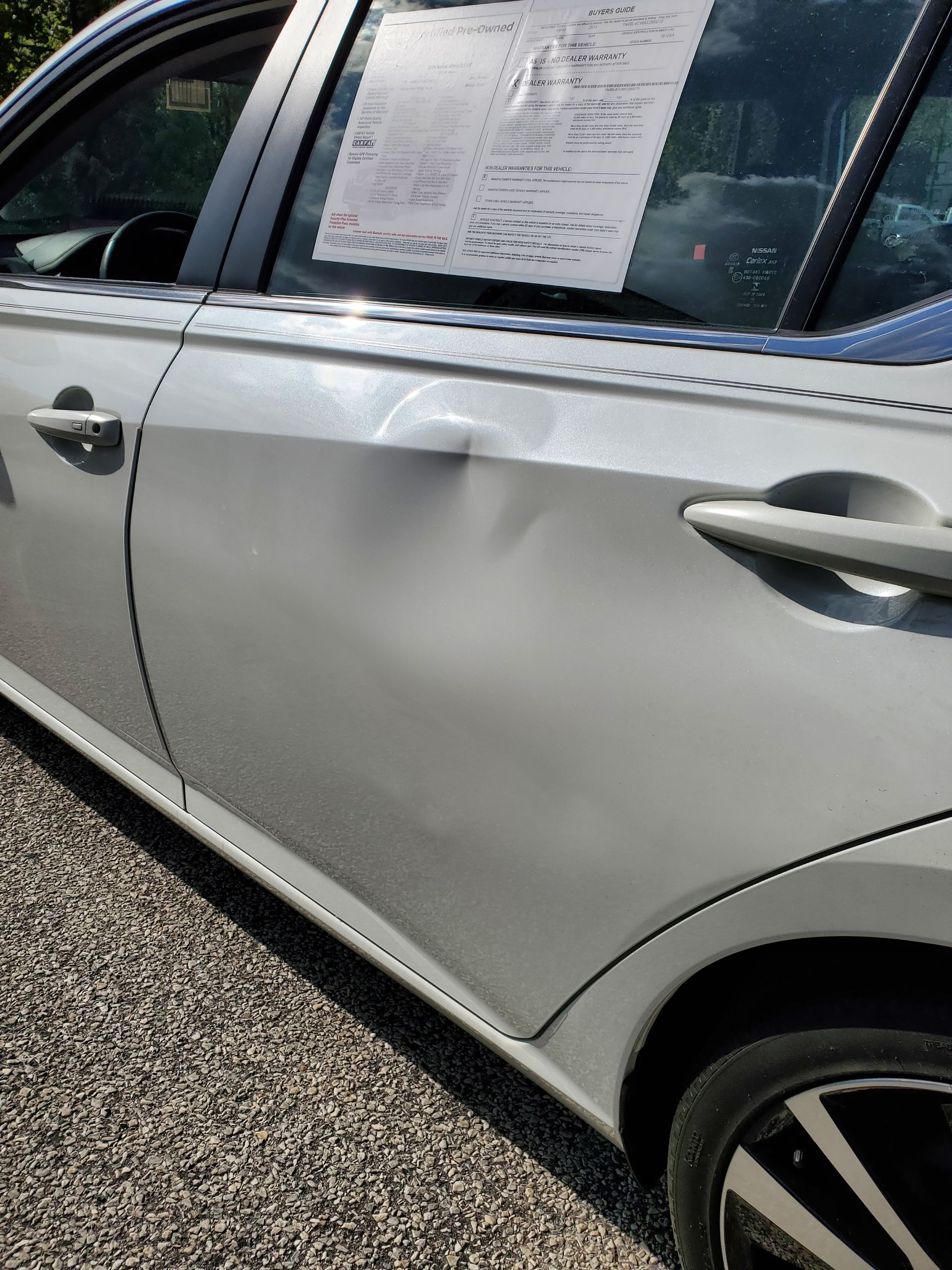White car side with a large dent on the rear door, parked outdoors with a sign in the window
