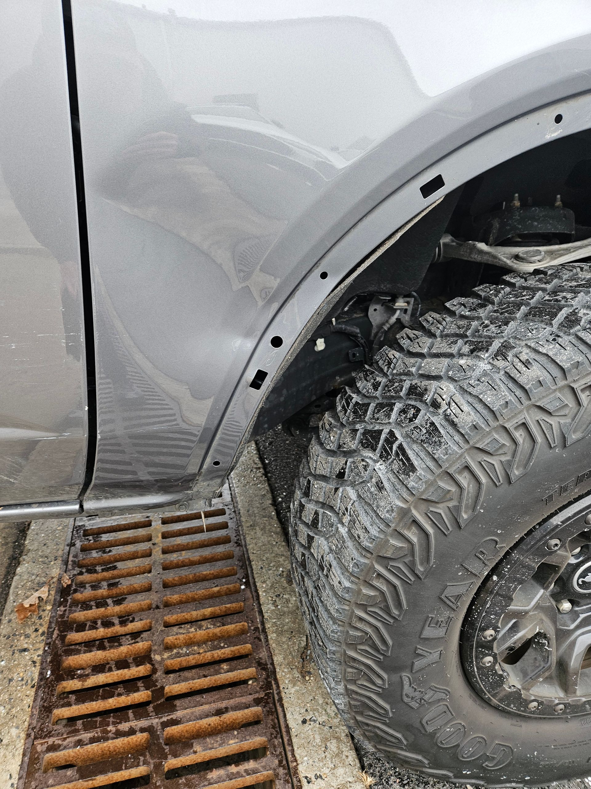 Close-up of a gray SUV’s front wheel and fender beside a rusty metal drainage grate.