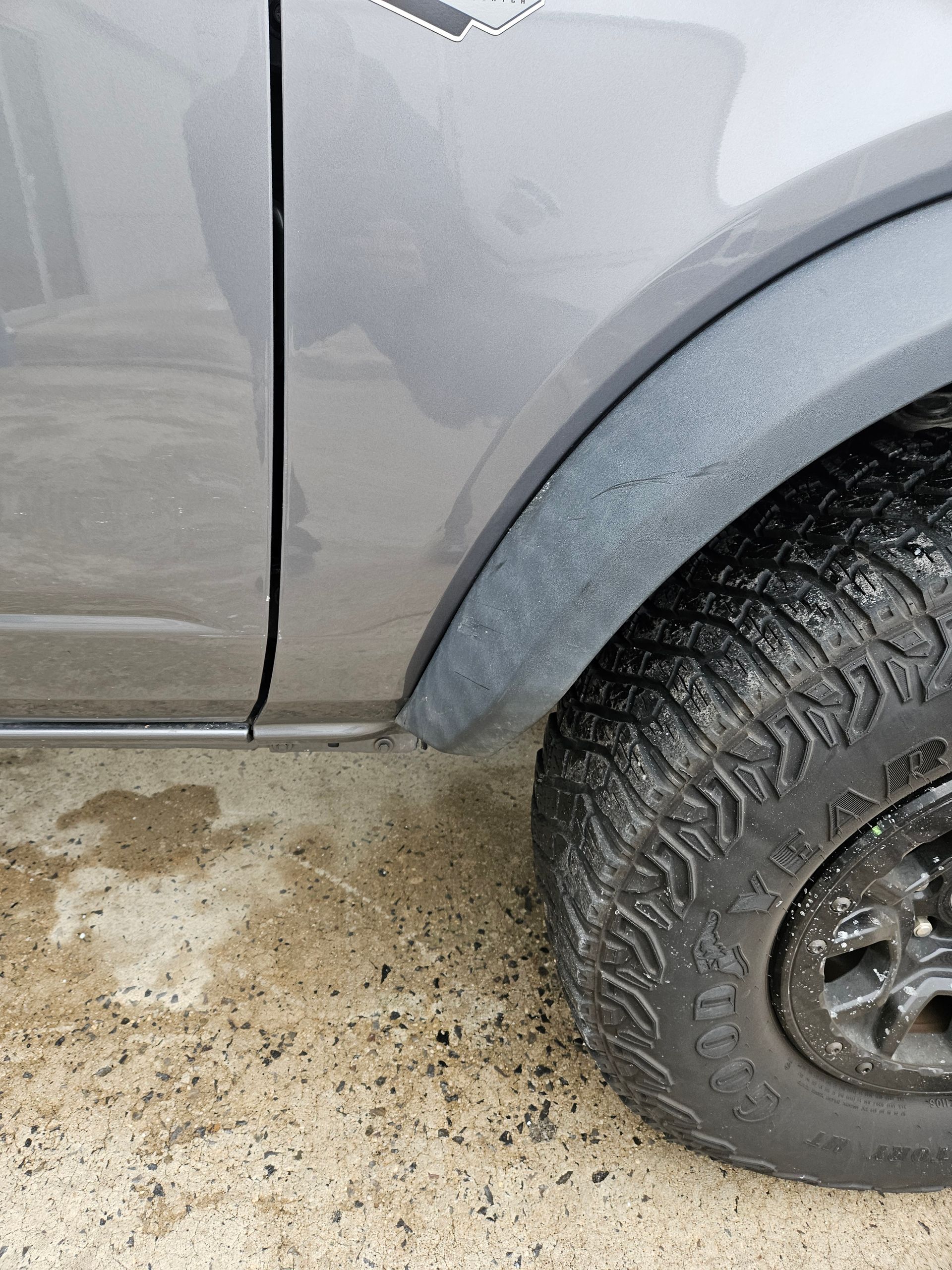 Close-up of a gray car’s side and wheel, with scuffed paint and muddy ground below.