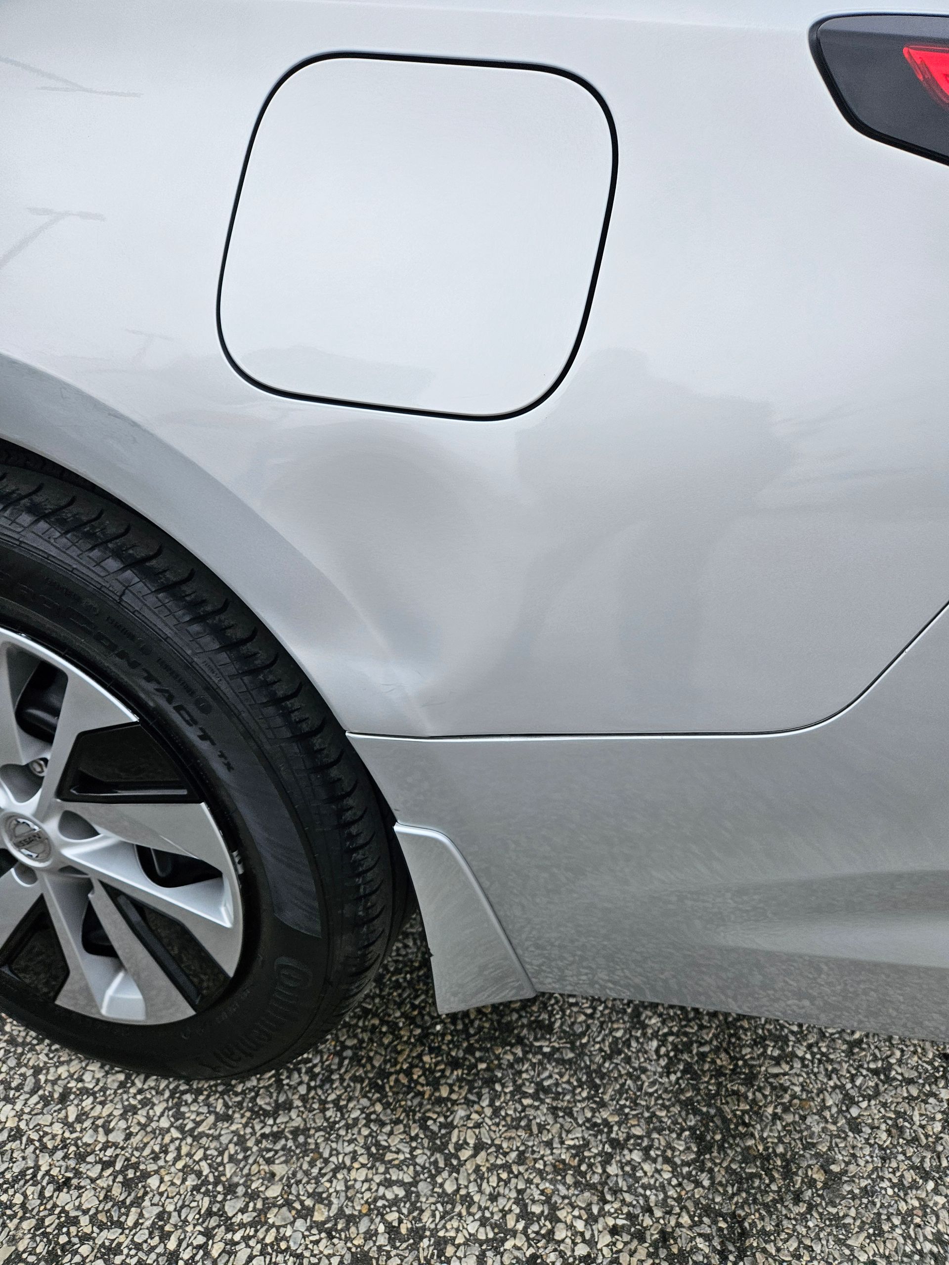 Close-up of a silver car’s rear side, showing the fuel door and back wheel on gravel.