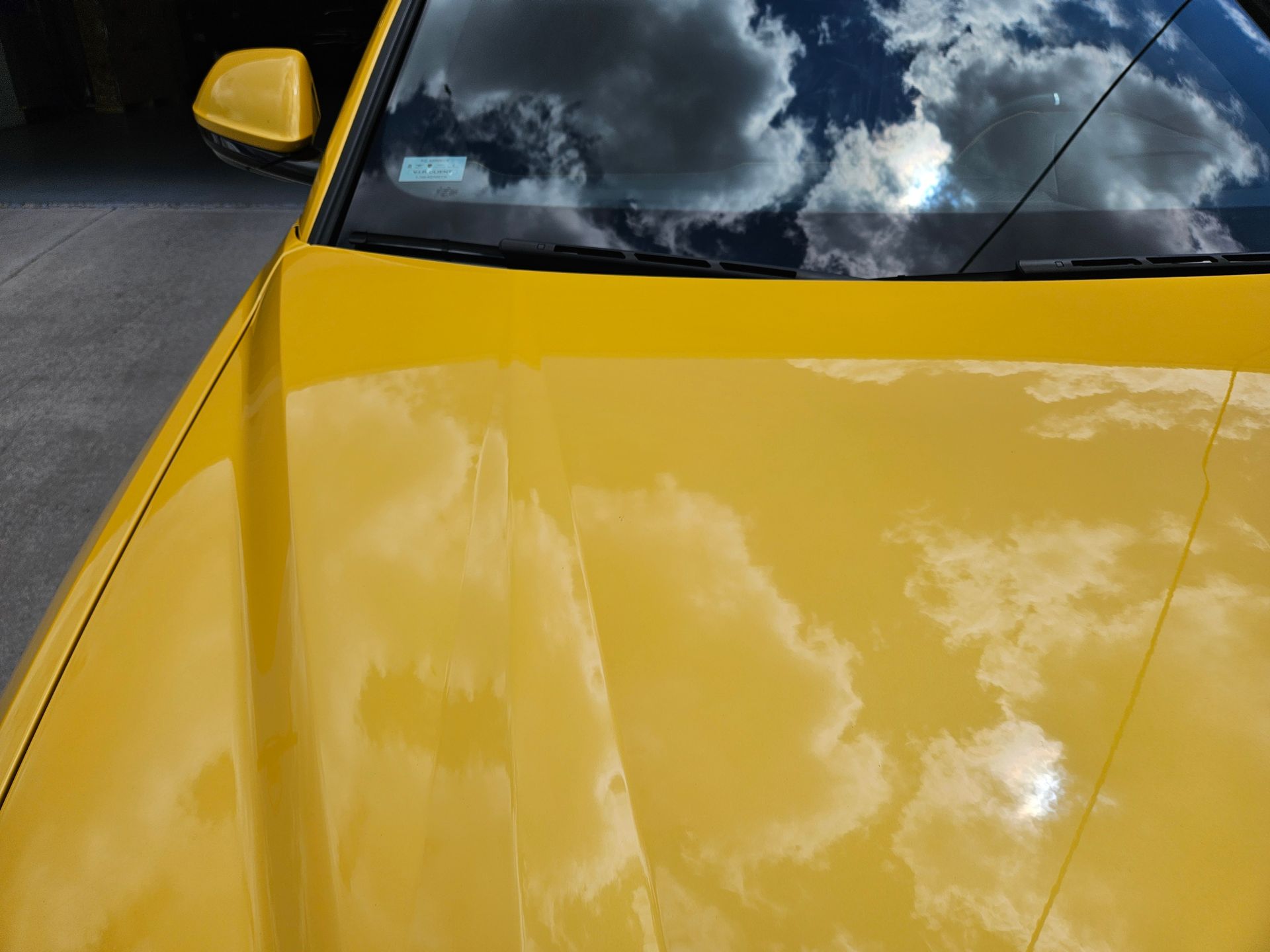 Close-up of a yellow car hood and windshield reflecting clouds, with a side mirror visible.