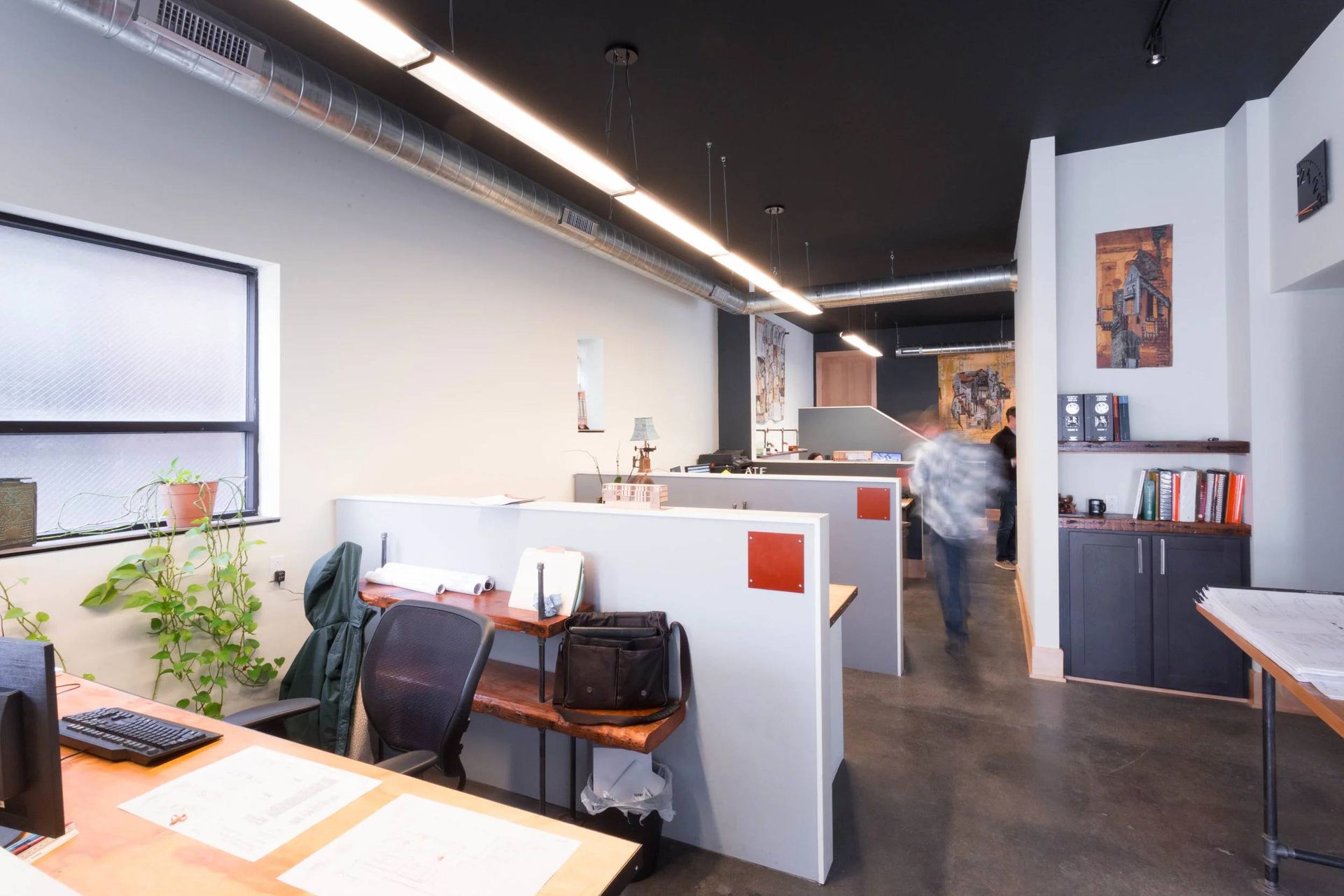 Open office interior with desks, chairs, plants, and a person walking down the hallway.